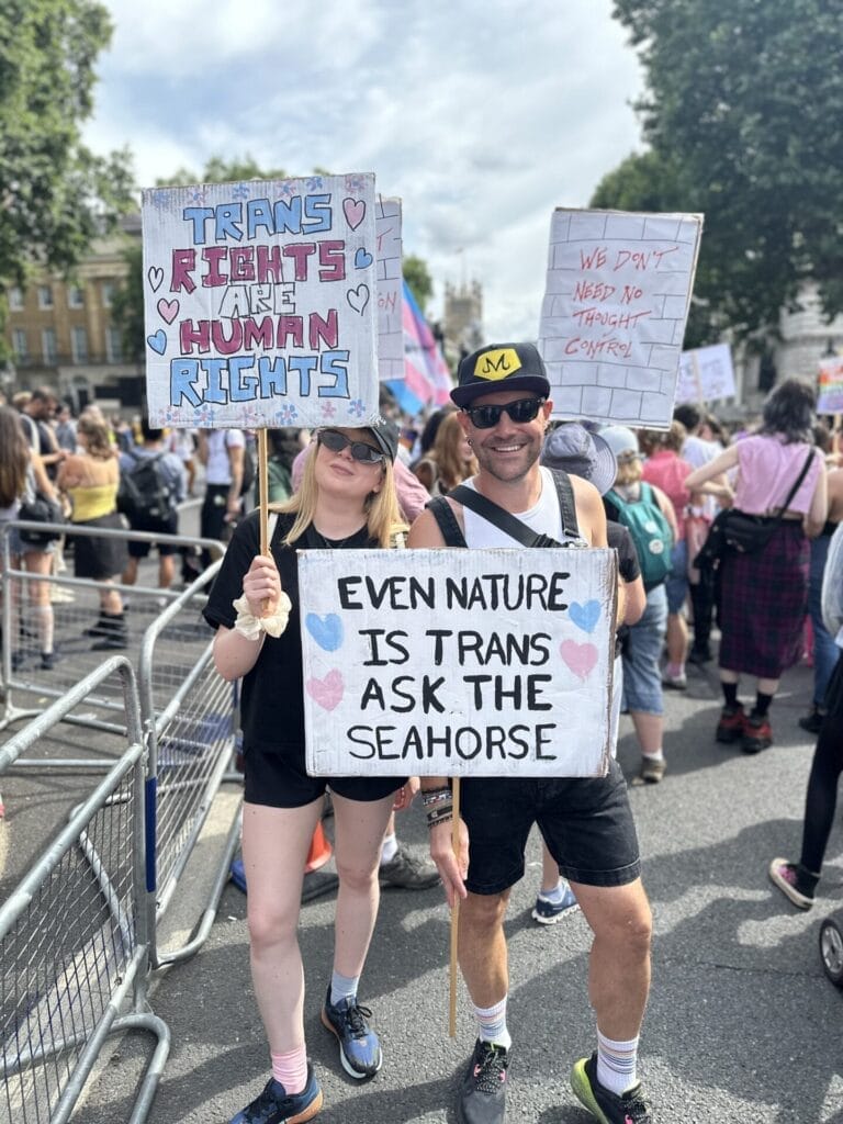 A man and a woman holding up signs during the march of the London Trans+ Pride 2025 with the woman's sign saying "Trans rights are human rights" and the man's sign says "Even nature is trans ask the seahorse".