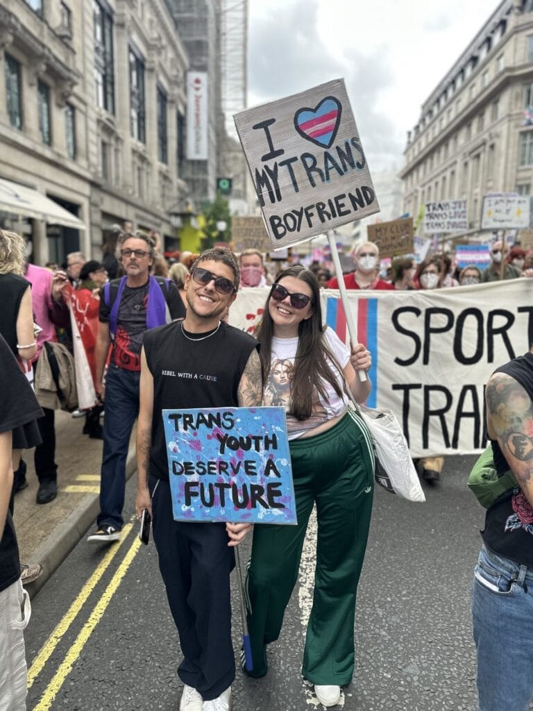 Two people during the march for London Trans+ Pride 2025, posing for a photo with the woman holding up a sign saying "I heart my trans boyfriend", and the man is holding a sign that says "Trans youth deserve a future".