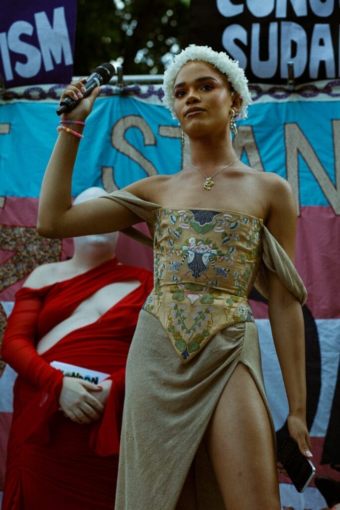 Yasmin Finney wearing a gold dress and holding a microphone, speaking at the London Trans+ Pride 2025 event.