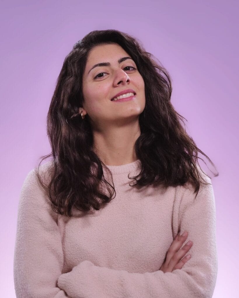 Official head shot of Paula Layoun who is smiling at the camera as her dark brown hair curls past her shoulders. She's wearing a pastel pink with beige tones jumper.