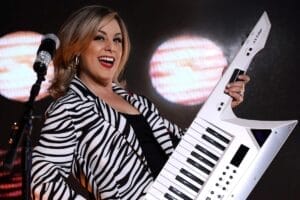 Valerie Rose performing on stage with her keytar, wearing a zebra-print jacket and a microphone on its stand in front of her. There's two big spotlights behind her too.