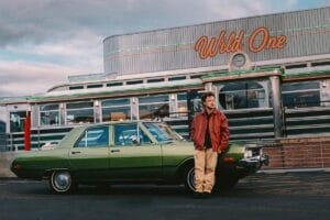 Cropped photo of Peech. standing outside Oaley Diner in a brown leather jacket, a black shirt and cream chinos, leaning against the bonnet of the green Dodge Dart getaway car.