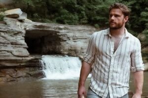 Callum Kerr posing in a white shirt with brown horizontal stripes, standing in front of a small waterfall with a pool of water. He's standing on a stone beach.