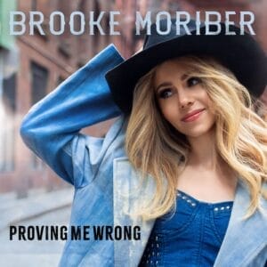 Official single cover artwork for "Proving Me Wrong" which sees Brooke Moriber wearing a light-blue jacket over a darker blue top and holding a black hat to her head with her right hand as her blonde hair falls down to her chest.