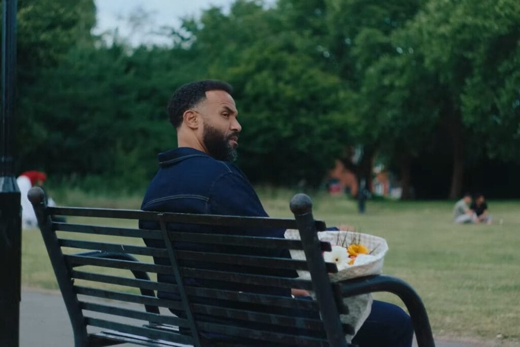 Screenshot from the "In It With You" music video which shows Craig David sitting at a black metal bench in a park, with a bouquet of flowers next to him.