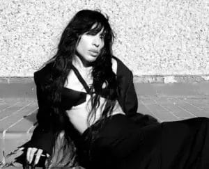 Black and white photo of Loreen wearing a jacket over a bra and a full-length skirt. She's sitting on the ground, leaning backwards and resting on her arms.