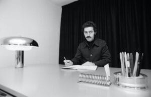 Black and white photo of Paco Rabanne sitting at his desk, writing in a book, with a metal lamp on the left and a pen pot with a note holder on the right.