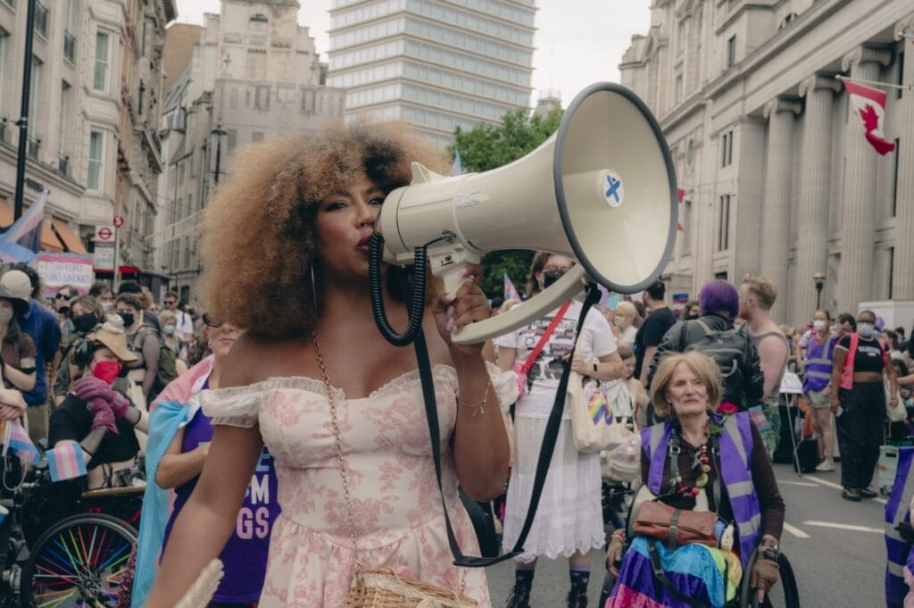 Olivia Campbell-Cavendish wearing a pink dress with her afro hair styled to perfection, speaking into a megaphone as part of the London Trans+ Pride 2025.