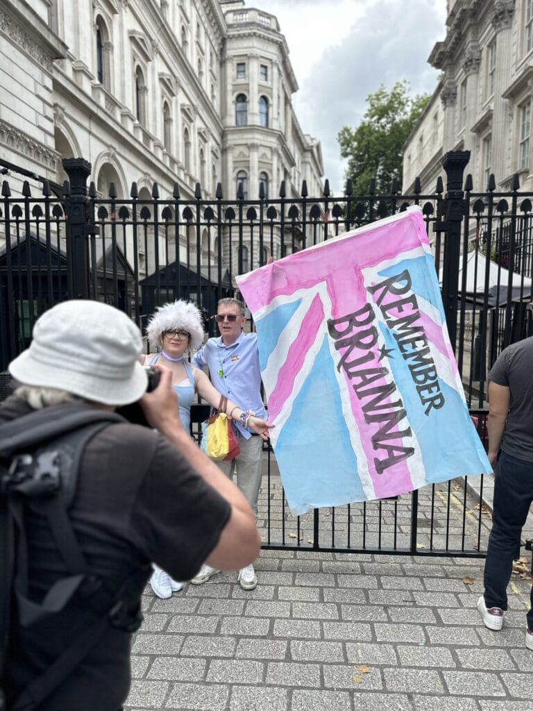 A man and woman holding up a flag that is in the trans colours that reads "Remember Brianna" in front of the Downing Street gates as part of the London Trans+ Pride 2025.