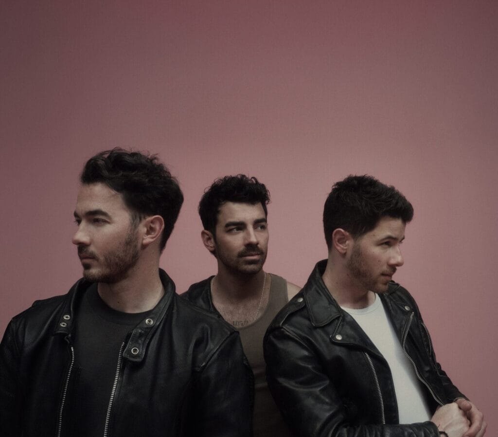 The Jonas Brothers posing together in leather jackets in front of a rose-pink background.
