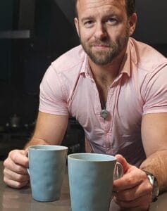 Mike Maimone posing in a pale-pink shirt holding the handles of two blue mugs of coffee that are sitting on a table.
