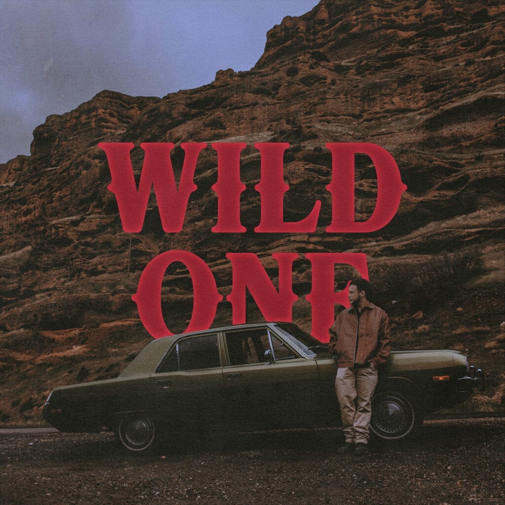 The official single artwork for "Wild One" which shows the title in big dark red lettering in front of a sand rockface with Peech. leaning against a green car, in front of the cliff face.