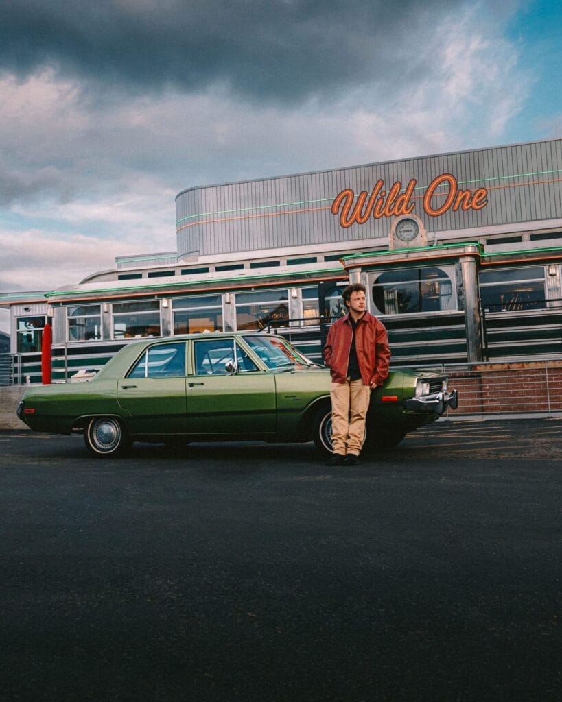 Full portrait photo of Peech. standing outside Oaley Diner in a brown leather jacket, a black shirt and cream chinos, leaning against the bonnet of the green Dodge Dart getaway car.