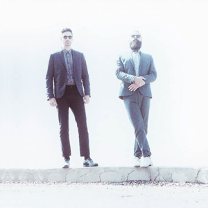 The duo known as Capital Cities standing on a wall both wearing suits. The image has an overexposure filter to it making it bright and ethereal.