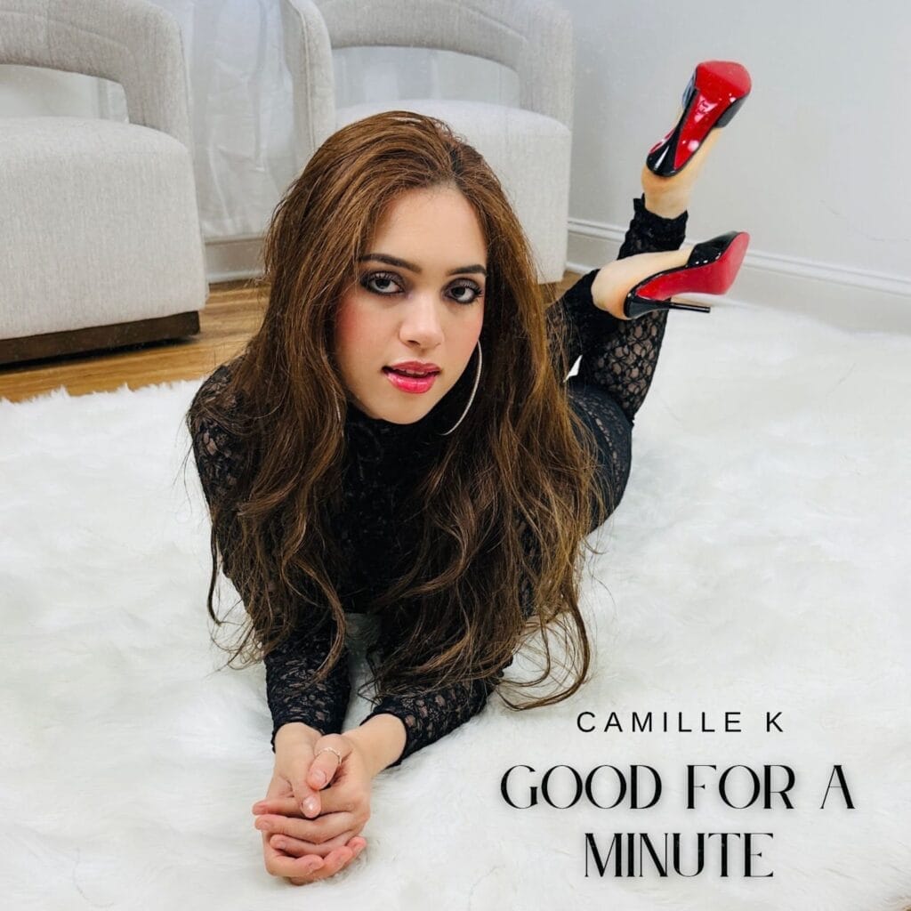 Official single cover artwork for "Good For A Minute", which sees Camille K wearing a black lace outfit and a pair of Louboutin heels. She's lying on her stomach with her knees bent and the heels in the air, leaning on her elbows with her hands out in front of her. The hardwood floor is covered by a grey-white rug which she is lying on. Her name and track's title are in black in the right-bottom corner.