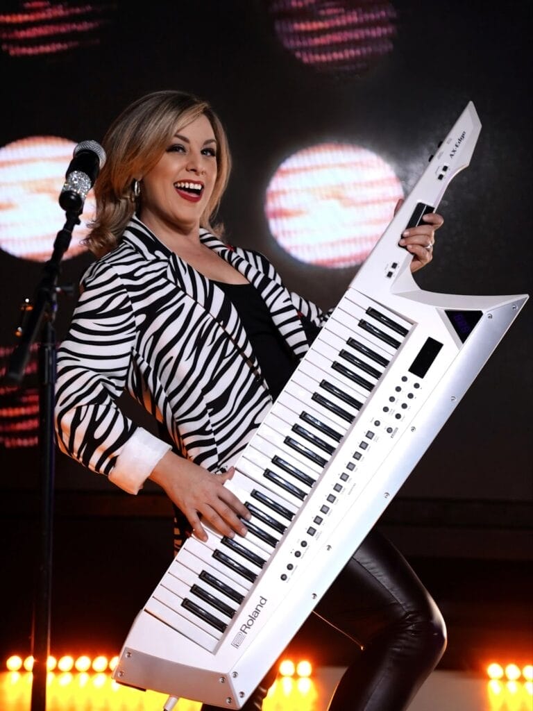 Full-length photo of Valerie Rose performing on stage with a silver keytar, wearing a zebra-print jacket, over a black top and a pair of black leggins, singing into a microphone that's in front of her, on a stand. There's two massive spotlights behind her.