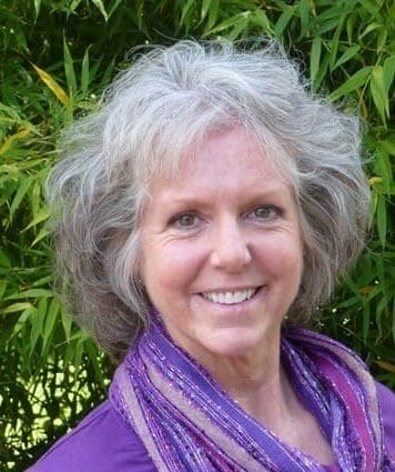 Headshot of Amanda Iles who is smiling at the camera and standing outside in front of some greenery. She's wearing a purple top and a matching purple scarf.