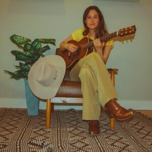 Sykamore dressed in yellow sitting in a chair with a guitar across her lap. A pale grey country-style hat is resting on the arm of the chair and there's a plant beside the chair.