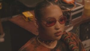 Still from the "Scorpion" official music video where Blake Red is sitting in the studio, wearing a tie-dye orange-to-black Korn t-shirt and orange shades.
