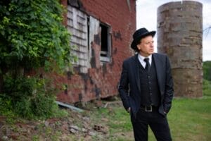 Steven Keene wearing a three-piece suit with a fedora hat standing on a patch of grass in front of a run down brick building.