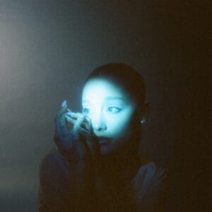 Ariana Grande posing with her hands clasped up to her cheek as a bright spotlight is shining on her face. The entire image has a blue filter to it.