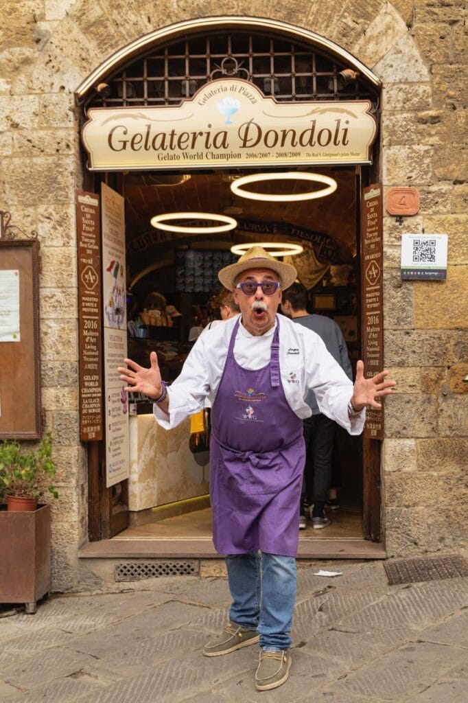 Sergio Dondoli with a shocked face outside Gelateria Dondoli, wearing a purple apron over a white shirt and jeans.