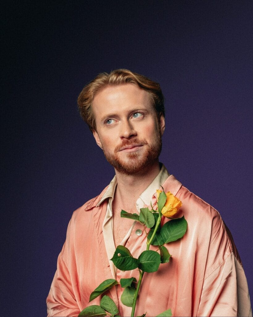 Interview: John Tothill on Malaria Trials, Burst Appendixes, and Finding Heaven at the Fringe 67 John Tothill giving a side-eye while wearing a pink silk-like shirt and holding a yellow rose to his chest backed by a dark blue background.