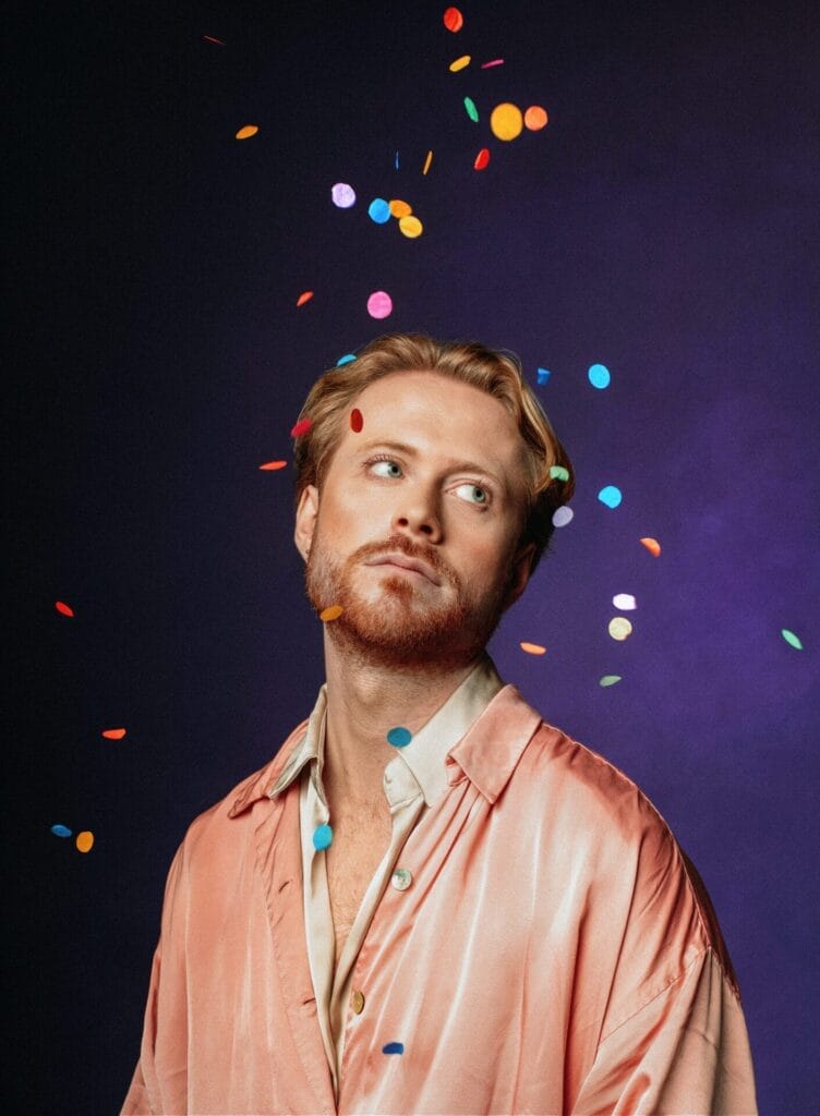John Tothill standing in front of a dark blue background while wearing a pink silk-like shirt as confetti rains down on him.