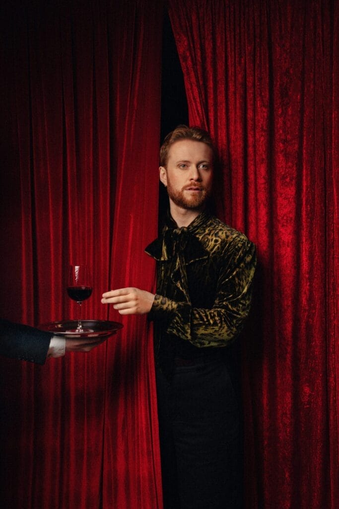 Interview: John Tothill on Malaria Trials, Burst Appendixes, and Finding Heaven at the Fringe 68 John Tothill peeking out of an opening in the red stage curtain, reaching for a glass of red wine which is standing on a serving trey and is being offered by someone off-screen on the left.