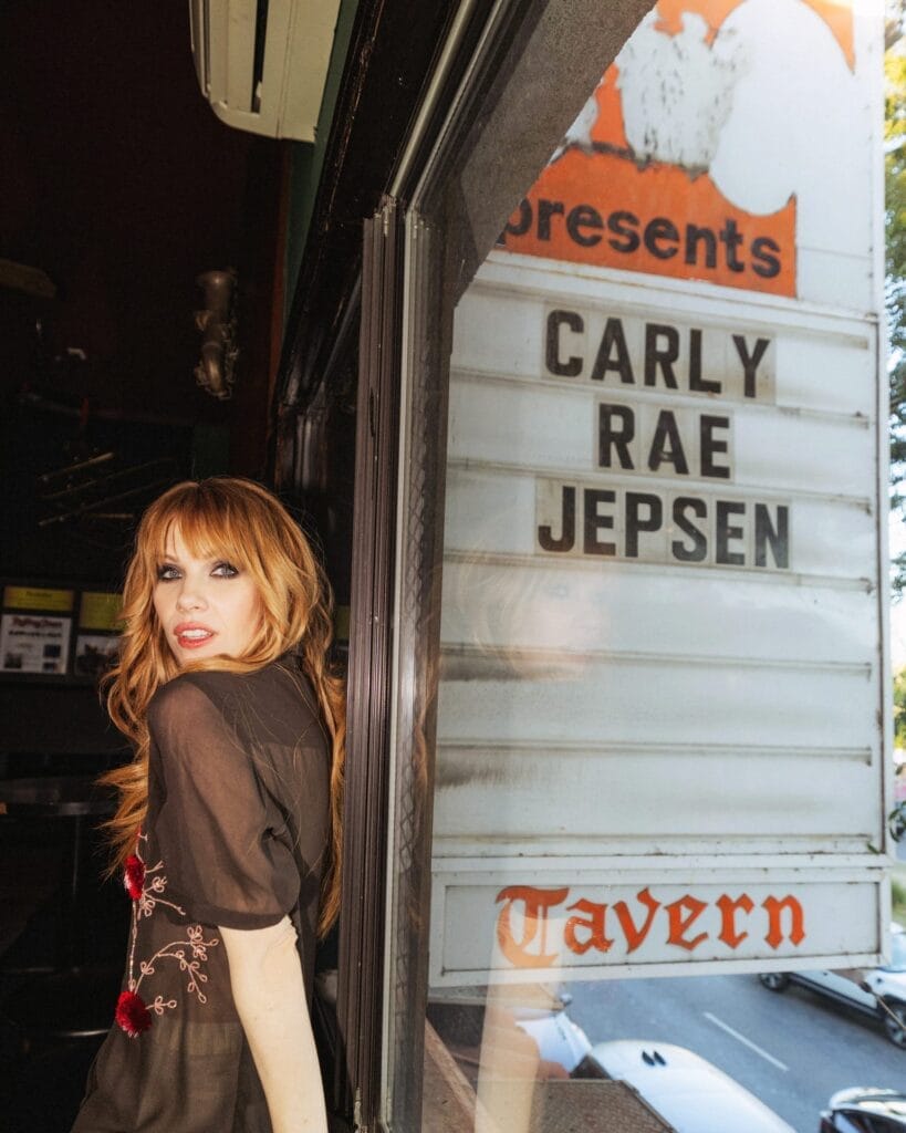 Carly Rae Jepsen Celebrates 10 Years of E•MO•TION With Special Anniversary Edition and New Single “More” 35 Carly Rae Jepsen posing outside The Troubadour in Los Angeles leaning against a glass plane which states her name outside. She's looking over her shoulder at the camera, wearing a black mesh dress embroidered with flowers.
