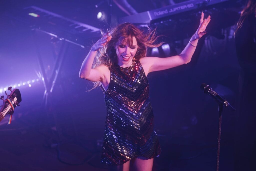 Carly Rae Jepsen performing on stage at The Troubadour in Los Angeles, wearing a rainbow sequin dress and she has her arms up in the air.