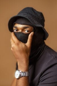 Professional head shot photo of TobySensey who is facing side-on and is wearing all black. He has a mask on that covers his entire head apart from his eyes, and he's holding his mask to his mouth. He also has a bucket hat on that is black, and a silver linked watch.
