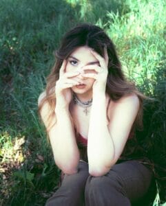 Hailey Hermida posing with her hands over her face and her elbows on her knees while sitting in some grass.