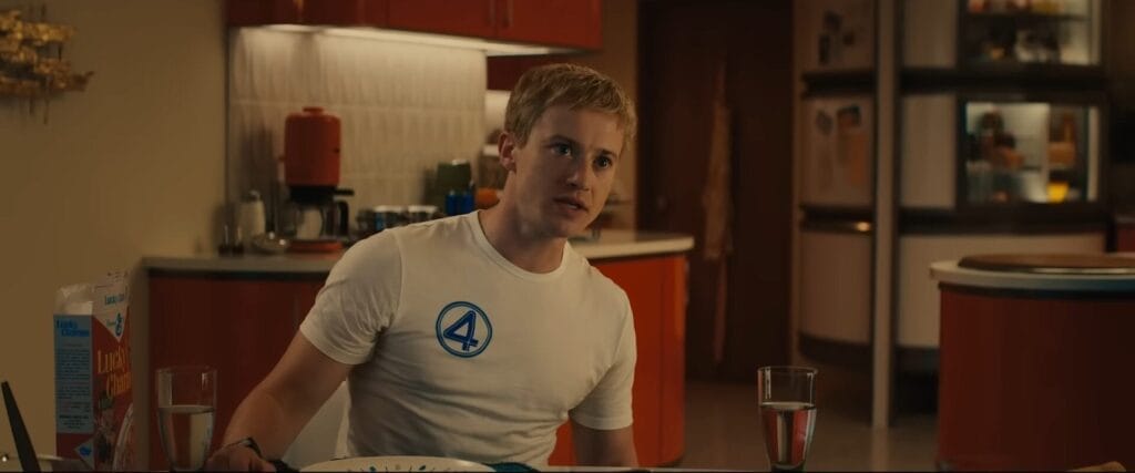 Still from the official trailer of The Fantastic Four: First Steps, which shows Johnny Storm sitting at the dining table with the kitchen behind him, with a bowl of cereal in front of him. He is wearing a tight white t-shirt with the Fantastic Four logo on it.