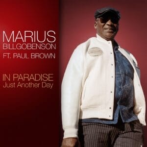 One of two official single cover artwork for "In Paradise – Just Another Day" which sees Marius Billgobenson wearing a white jacket over a blue shirt and paired with black pin-striped trousers. He is also wearing shades and a black flat cap. Behind him is a red background with text of the artist name and song title on the left.