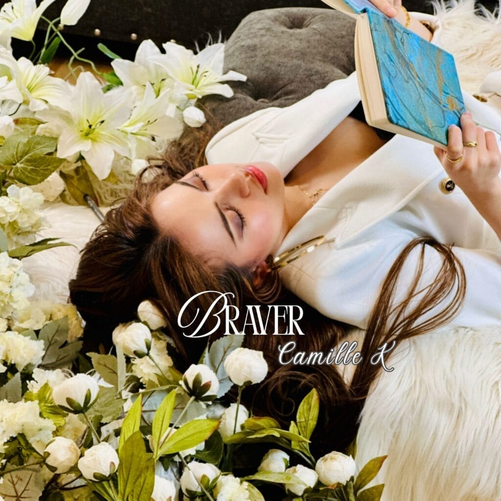 Camille K Explores Vulnerability and Power in New Singles “Braver” and “Facade” 26 Official single cover artwork for "Braver" which shows Camille K lying on her back surrounded by white flowers in what looks like a field. She's wearing a white jacket and is reading a book.