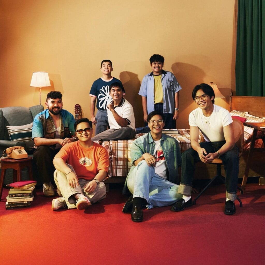 Seven-piece band, Lola Amour, are in a cosy room with warm lighting. They smile casually, sitting and standing around a bed, creating a relaxed, friendly atmosphere.