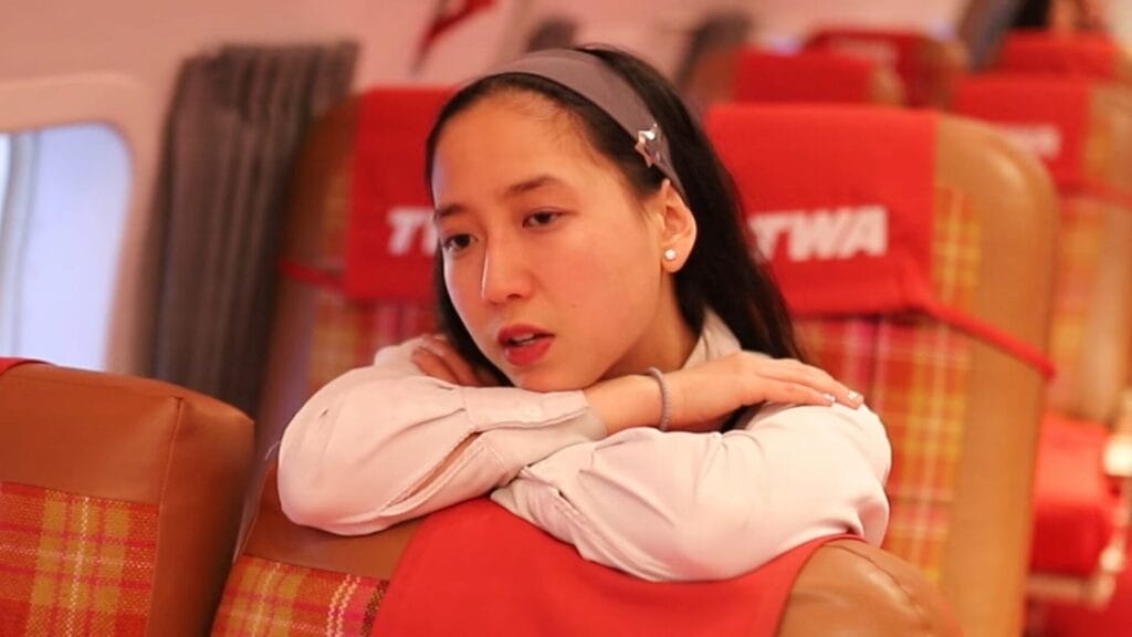 Still from "Gut Feeling" music video which shows ÊMIA with a grey headband resting her arms on a plaid airplane seatback, appearing thoughtful. The vintage cabin has red TWA-branded headrest covers.