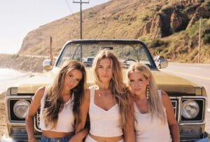 The girl group Just Jayne posing together in white tops in front of a car with a big bonnet / hood, on an open road with a hill and a telegraph pole behind them.