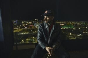 ZAYN sitting in front of a cityscape at night with the city lit up in bright lights, as he wears black clothing, a leather jacket, and a Found black baseball cap.