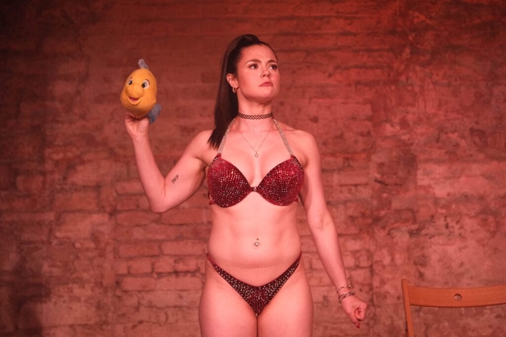 Megan Prescott on stage performing her one-woman show Really Good Exposure, wearing a red sparkly bra and knickers and holding up a plush of Flounder from The Little Mermaid in her right hand.