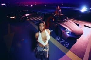Still from the music video of "S.O.S." which sees Antonia in a car park wearing a white cleavage-showing vest-like top with jeans, dancing in front of a Porsche. Her two back-up dancers are in the car.