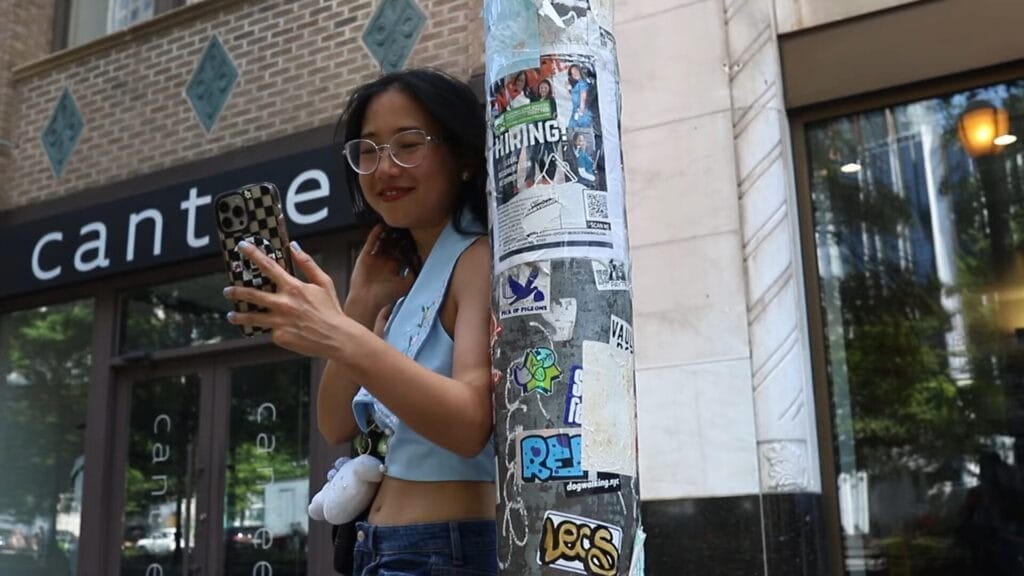 Still from "The Plot" music video which shows ÊMIA smiling while wearing glasses and a blue top leaning against a sticker-covered pole, holding a smartphone. She's standing outside a building conveying a casual and urban vibe.