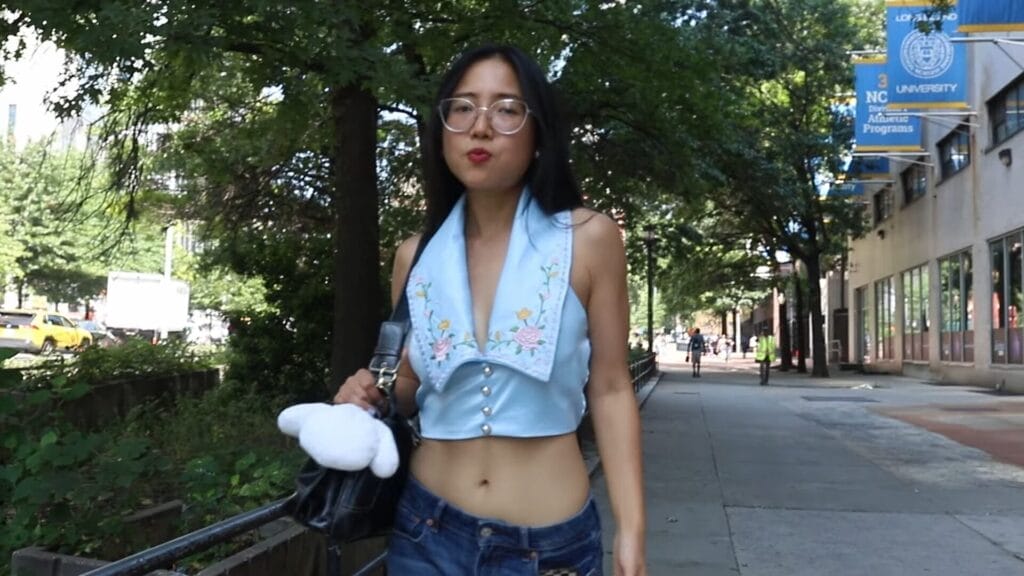 Still from "The Plot" music video which shows ÊMIA walking on a sunlit city sidewalk, wearing a pair of glasses, a light blue floral top and jeans, carrying a purse with a plush toy. The background shows trees and buildings.