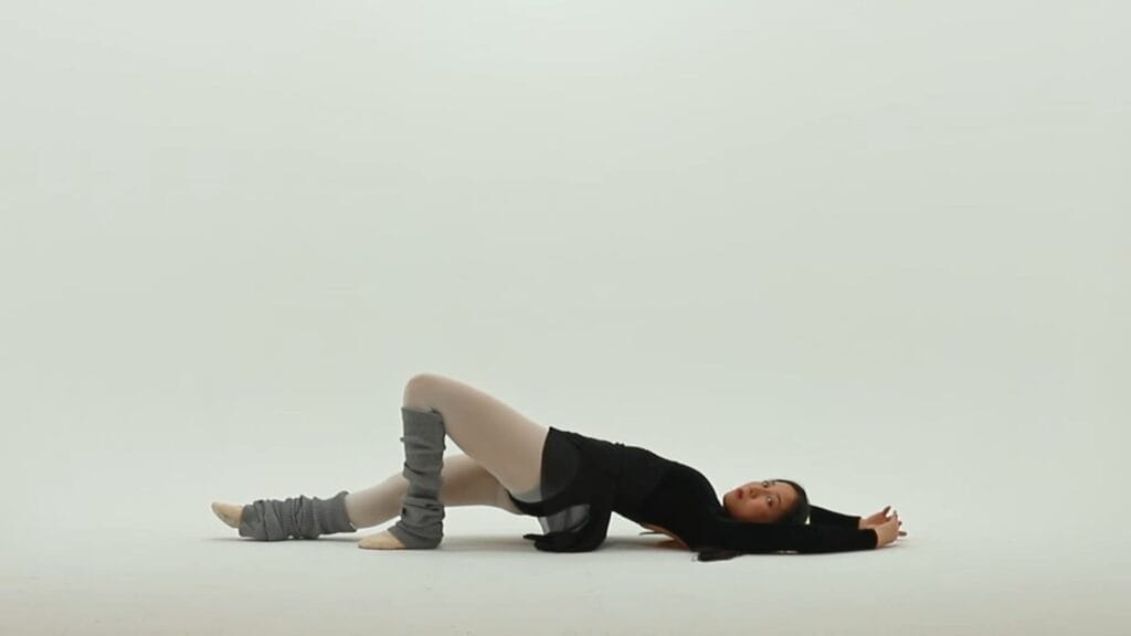 Still from "Gut Feeling" music video which shows ÊMIA looking like a ballet dancer in a black leotard and gray leg warmers posing gracefully on the floor. The serene, minimal background enhances the elegance of the pose.