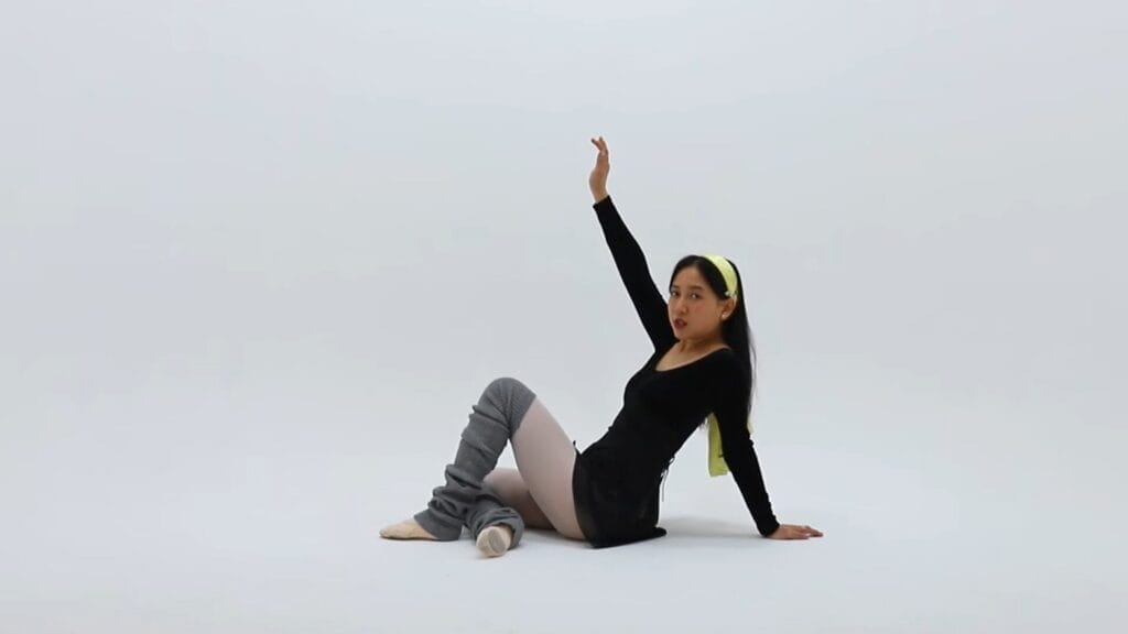 Still from "Gut Feeling" music video which shows ÊMIA in a black leotard and grey leg warmers sitting on the floor against a white background, one arm raised, conveying a poised, dance-like posture.