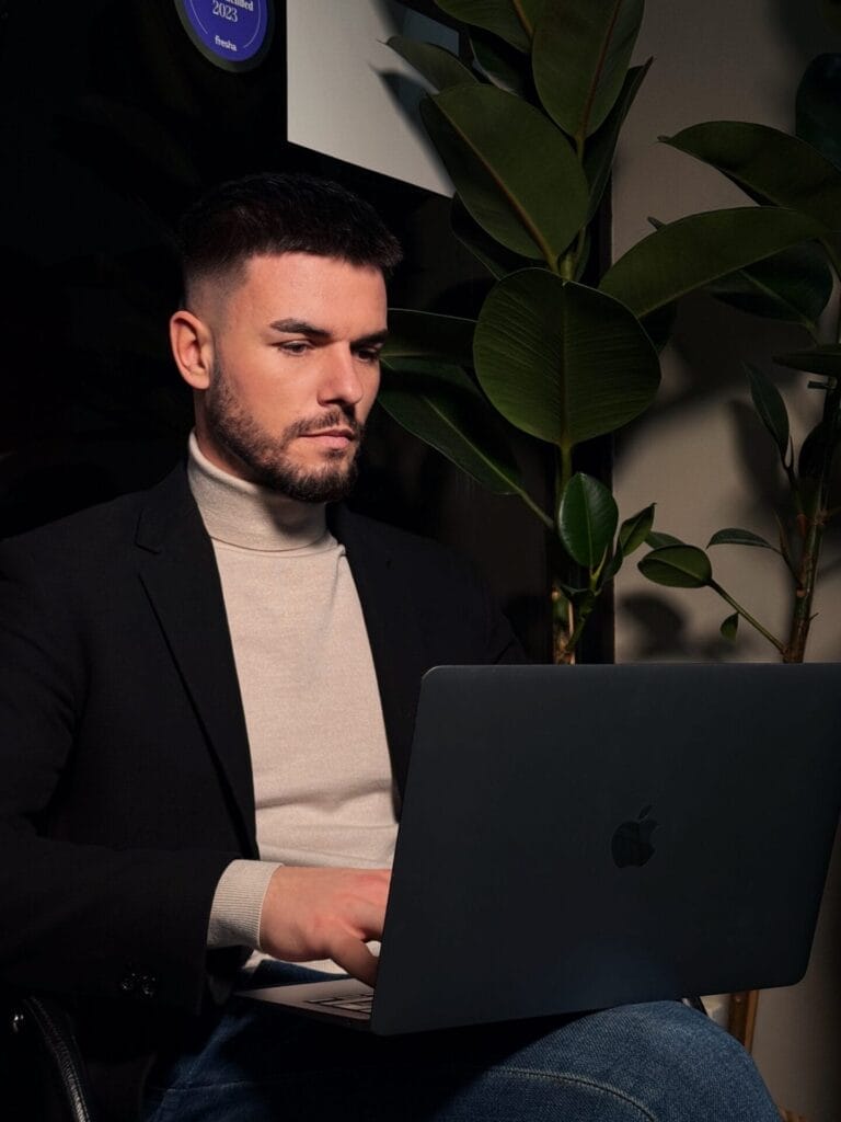 “Haters Are Free Publicity”: Ukrainian Singer Ihor Tselyp Opens Up About Turning Negativity Into Creative Power 15 Ihor Tselyp with a beard, wearing a blazer and a cream turtleneck, working attentively on a laptop. He's seated near large green plants, creating a focused and calm atmosphere.