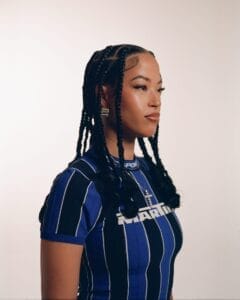 Press photo of Jazzy who is turned slightly to the right, and is wearing a black and blue horizontal-striped football-like top with a cross necklace.