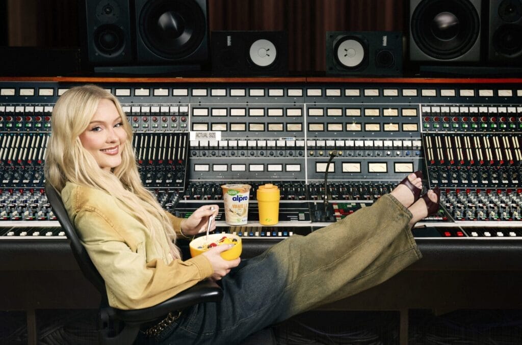 Zara Larsson relaxing with feet up on a mixing console in a studio, holding a bowl of cereal with Alpro yogurt. Studio equipment surrounds her, creating a casual, playful vibe.