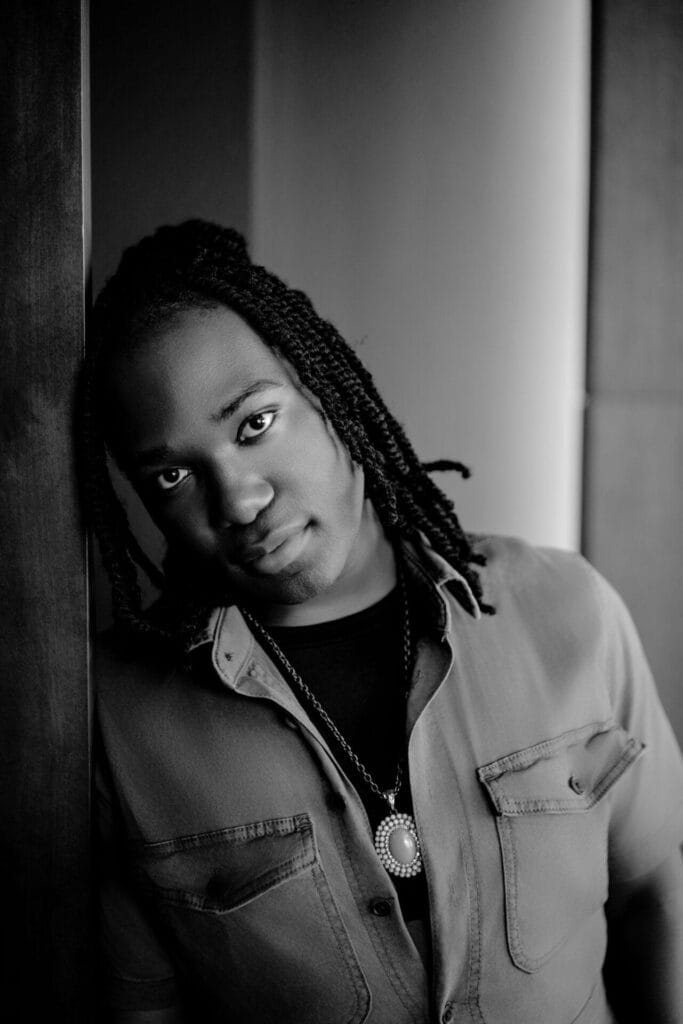 Black and white portrait of Jourdan Frost with braided hair, leaning against a wall. They wear a buttoned shirt and necklace, looking thoughtfully at the camera.